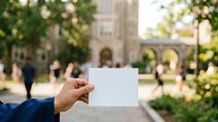Cropped hand holding blank white card outdoors near historic stone building with green trees and blurred people in background. Use for business announcements, event promotions, educational messaging.