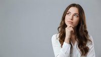 Young Caucasian businesswoman in white shirt thinking with hand on chin, looking up thoughtfully against gray background for corporate concepts.