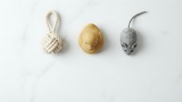 Three pet toys arranged on marble surface: rope knot ball, yellow rubber duck, and gray felt mouse for cats and dogs.
