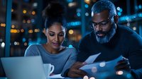 African American business colleagues reviewing documents and working on laptop during late evening meeting with city lights and bokeh background.