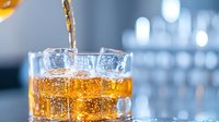 Amber liquid being poured into glass with ice cubes, creating dynamic splash and bubbles against blurred bar background. Golden beverage catching light with refreshing appeal.
