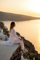 Pregnant woman in white clothing sitting on stone wall overlooking golden sunset ocean view from Mediterranean cliff village for maternity lifestyle.
