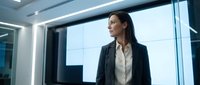 Professional businesswoman in dark suit standing by office windows looking thoughtful in modern corporate building interior with city view.