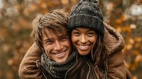 Happy interracial couple embracing outdoors in autumn wearing cozy winter clothing with warm smiles and natural bokeh background.