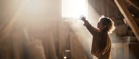 Young woman with arms raised in sunlit attic space expressing freedom and joy in warm golden light streaming through window.