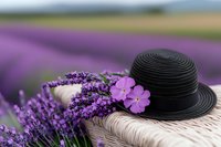 Black straw hat decorated with fresh lavender flowers and pink phlox on wooden surface against blurred purple lavender field background, romantic summer mood.