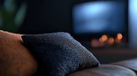 Cozy brown leather sofa with dark blue throw pillow in dimly lit living room. Blurred fireplace with warm glowing flames creates relaxing atmosphere for home decor.