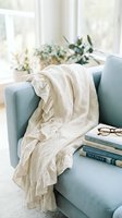 Cozy reading nook with soft cream throw blanket draped over light blue armchair, stacked books, eyeglasses, and fresh flowers by bright window for home decor and lifestyle content.