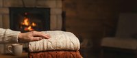 Cozy autumn scene with folded knit sweaters and a hand resting on them near a fireplace with warm flames glowing in the background, perfect for fall and winter lifestyle content.