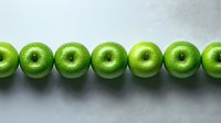 Fresh green apples arranged in straight line on light gray background with water drops, showing natural texture and vibrant color. Minimal food photography.