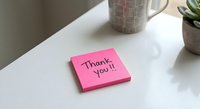 Pink sticky note with thank you message on white desk surface next to coffee mug and succulent plant for office gratitude and appreciation concepts.