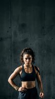 Fit woman athlete in black sports bra and shorts standing confidently against dark textured wall, ready for workout or training session.