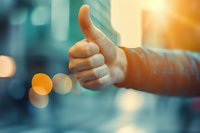 Close-up of hand showing thumbs up gesture against blurred urban background with warm sunset bokeh lights, expressing positivity and approval.