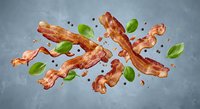 Crispy bacon strips flying with fresh basil leaves and black peppercorns on gray background. Breakfast meat ingredients in motion for culinary designs.