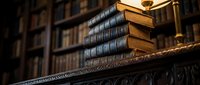 Antique leather-bound books stacked on ornate wooden shelf in classic library setting with warm lighting and blurred bookcase background.
