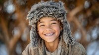 Happy mixed race child wearing fur winter hat with ear flaps smiling with dental braces in snowy forest setting for seasonal lifestyle content.