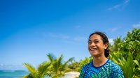 Young woman with dark hair and Southeast Asian ethnicity smiling outdoors on tropical beach with palm trees and turquoise ocean water, wearing blue patterned shirt for travel, vacation, lifestyle content.