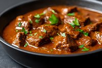 Tender beef chunks in rich red curry sauce garnished with fresh herbs, served in black ceramic pan on dark background. Traditional spicy meat stew.