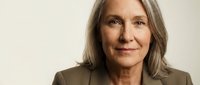 Mature woman with long gray hair wearing olive blazer, professional headshot on neutral background for corporate and business use.
