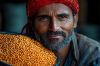 Middle-aged South Asian farmer with gray beard wearing red headscarf smiles while holding large bowl of fresh harvested orange lentils, representing agriculture and farming.