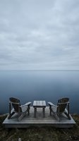 Wooden Adirondack chairs and table on a deck overlooking calm blue ocean waters and cloudy sky, perfect for relaxation and outdoor seating design projects.