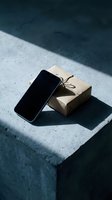Smartphone with blank screen leaning against kraft paper gift box with twine on concrete surface in natural sunlight, suitable for e-commerce and technology marketing.