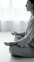 Woman in linen clothing meditating in lotus position by bright window, practicing mindfulness and yoga for wellness and mental health.