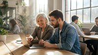 Senior woman with white hair and younger bearded man of Middle Eastern descent reviewing laptop together in bright office workspace with colleagues and plants in background.