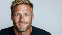 Confident mature Caucasian man with stubble beard and blonde spiky hair wearing black t-shirt smiling at camera against light background, head and shoulders portrait.