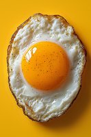 Sunny side up fried egg with crispy edges and bright orange yolk sprinkled with black pepper on vibrant yellow background, macro food photography.