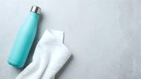 Turquoise insulated water bottle with stainless steel cap next to white towel on gray textured surface, ideal for hydration and wellness product marketing.