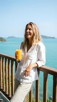 Woman with blonde hair, Caucasian ethnicity, wearing white linen shirt and pants, holding glass of orange juice on tropical beach balcony overlooking turquoise ocean. Summer vacation lifestyle.
