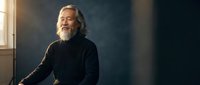 Senior Caucasian man with gray beard in black sweater sitting thoughtfully by window with warm natural light creating atmospheric portrait for lifestyle content.