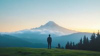 Silhouette of lone person standing on grassy hill overlooking majestic mountain peak at sunset, evergreen forest in foreground creates serene wilderness landscape.
