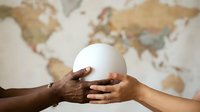Two hands of different skin tones holding a white globe together against a blurred world map background, symbolizing global unity, cooperation, and international partnership for business and peace.