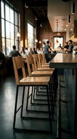 Row of wooden bar stools with black metal frames at counter in modern industrial restaurant or cafe with blurred guests dining in background and large windows.