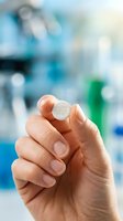 Hand holding round placebo pill in laboratory setting with blurred scientific equipment and bottles in background, suitable for pharmaceutical, medical research, clinical trial, and healthcare marketing materials.