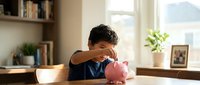 Young boy saving money in pink piggy bank at home desk with books and plants in bright natural light for financial education concept.