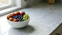 Assorted fresh berries and sliced kiwi fruit arranged in white bowl on marble countertop near window, ready for healthy breakfast or dessert preparation.