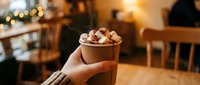 Hand holding a steaming paper cup of hot chocolate topped with toasted marshmallows in a cozy warm cafe with bokeh lights.