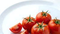 Fresh red tomatoes with green stems on white plate, one sliced showing interior, healthy organic vegetables for cooking and nutrition concepts.