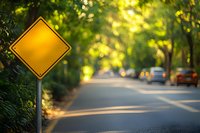 Empty yellow diamond warning sign on blurred suburban road with soft bokeh sunlight filtering through trees and parked cars in background.