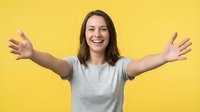 Young woman with long dark hair and fair complexion wearing light gray t-shirt, arms outstretched in celebratory gesture against bright yellow background, expressing joy and enthusiasm.