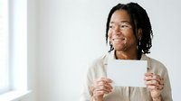 Young African American woman with long braided hair wearing a beige cardigan, holding blank white card, smiling while looking away in bright studio setting.