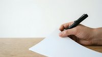 Close-up of hand holding pen over blank white paper on wooden desk, ready to write or sign document for business, education, or creative work.