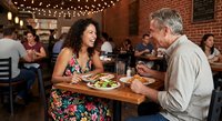 Mature couple enjoying romantic dinner date at cozy restaurant with brick walls and string lights creating intimate dining atmosphere.