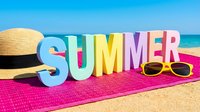 Colorful SUMMER letters on pink towel at sandy beach with straw hat and yellow sunglasses, bright blue sky and sea in background.