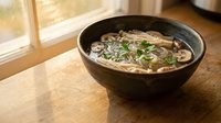 Steaming bowl of Asian noodle soup with mushrooms and green onions in dark ceramic bowl on wooden table near sunny window for restaurant menu design.