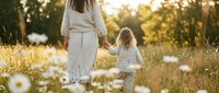 Mother and daughter walking hand in hand through wildflower meadow during golden hour sunset, family bonding time in nature with warm sunlight filtering through trees.