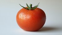 Fresh ripe tomato with water drops on red skin and green stem against light background, macro food photography showing natural texture and freshness.